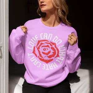 May include: A light purple sweatshirt with a white graphic of a rose and the text "What I Can't Do, Love Can Do".