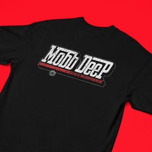 May include: Black t-shirt featuring the text "Mobb Deep" in a white, distressed font with a red accent. The text "QUEENSBRIDGE" is below the main text. The shirt has a crew neck and short sleeves.