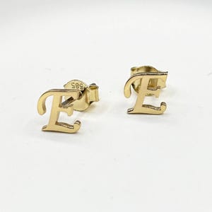 May include: A pair of gold-colored stud earrings, each shaped as the letter "E". The earrings are displayed on a white background. The post of one earring is visible, with the number "385" stamped on it.