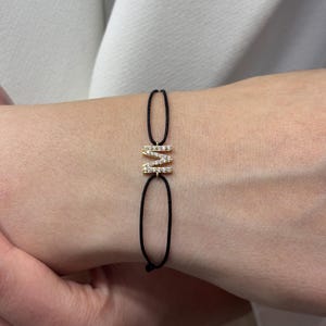 Puede incluir: Una pulsera dorada con la letra "M" incrustada con pequeñas piedras transparentes. La letra está unida a una pulsera de cordón negro. La pulsera se muestra en la muñeca de una persona.
