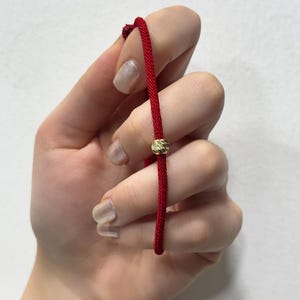 Puede incluir: Una pulsera de cordón rojo con una cuenta dorada. La pulsera se sostiene sobre un fondo blanco. El cordón es de color rojo vibrante y la cuenta tiene una superficie texturizada.