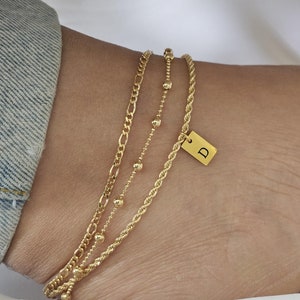 May include: Three gold anklets with a charm that says "D" on the ankle. The anklets are layered on top of each other. The anklet on the bottom is a thick chain, the middle anklet is a thin chain with small beads, and the top anklet is a rope chain.
