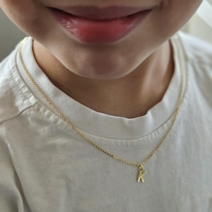May include: A gold-colored necklace with a small letter "K" pendant. The necklace is worn on a person wearing a white t-shirt. The chain is delicate and the pendant is a simple, elegant design.