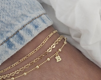 May include: Three gold anklets layered on an ankle. The top anklet is a simple chain, the middle anklet is a chain with heart-shaped links, and the bottom anklet is a beaded chain with a charm that says "B".