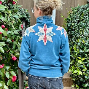 May include: A blue sweatshirt with a starburst design on the back, worn with dark wash jeans and black trainers. The starburst design has white, red, and blue accents. The person is standing on concrete steps with greenery on either side.