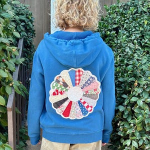 May include: A blue hoodie with a colorful, circular patchwork design on the back. The patchwork features various fabric patterns and colors, including red, white, and blue. The person is wearing khaki pants and green sandals.