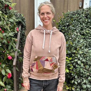 Puede incluir: Una mujer con una sudadera con capucha marr&oacute;n claro con un bolsillo de retazos, combinada con vaqueros grises oscuros y zapatos sin cordones negros. La sudadera tiene una capucha con cord&oacute;n y los vaqueros est&aacute;n doblados. Est&aacute; de pie frente a una puerta y arbustos verdes.