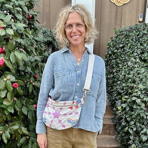 Puede incluir: Una persona con una camisa azul claro, pantalones color caqui y sandalias plateadas. Lleva un bolso cruzado con correa blanca y un diseño de patchwork con formas rosas, moradas y azules. El fondo incluye arbustos verdes y una puerta de madera.