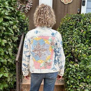 Puede incluir: Una chaqueta de mezclilla de lavado claro con un patrón de teñido anudado azul y un gran parche acolchado rectangular multicolor en la espalda con estampados florales y geométricos en amarillo, rosa y azul, junto con jeans azules de corte recto.