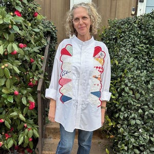 May include: A white button-down shirt with colorful patchwork details on the front and sleeves. The shirt is paired with blue jeans and dark ankle boots. The outfit is worn outdoors, in front of a green bush.