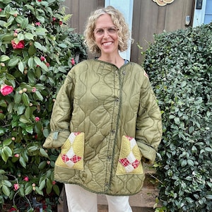 May include: Olive green quilted jacket with a diamond pattern, featuring two patch pockets with yellow and red plaid accents. The jacket has a button-up front and is worn with off-white pants and black sandals.