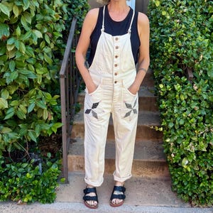 Può includere: Salopette in denim bianco sporco con chiusura frontale a bottoni, due tasche anteriori con dettagli patchwork a forma di stella scura e orli risvoltati. La salopette è indossata sopra una canotta nera senza maniche con sandali neri a due fasce.