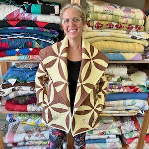 May include: A woman wearing a brown and cream-colored quilted jacket with a floral pattern. The jacket is open, revealing a black top. Shelves behind her are stacked with various colorful quilts, showcasing a collection of handmade textiles.