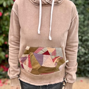 Puede incluir: Una sudadera con capucha color canela con un bolsillo de retazos con varios colores y texturas. La sudadera tiene una capucha con cord&oacute;n y pu&ntilde;os acanalados. La prenda la lleva una persona.