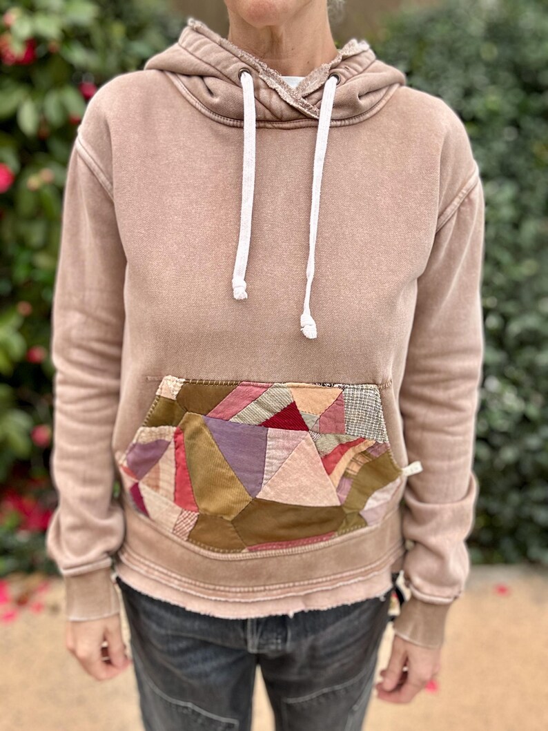 Puede incluir: Una sudadera con capucha color canela con un bolsillo de retazos con varios colores y texturas. La sudadera tiene una capucha con cord&oacute;n y pu&ntilde;os acanalados. La prenda la lleva una persona.