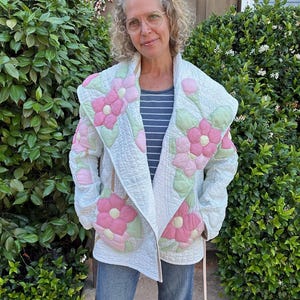 May include: A quilted jacket in white featuring large, appliquéd pink flowers with light green leaves. The garment has a wide shawl collar, long sleeves, and a textured, stitched pattern throughout the fabric. It is worn over a striped shirt.