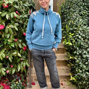 Puede incluir: Una persona con una sudadera azul con cordones blancos y cuello alto, combinada con vaqueros negros y zapatos negros. La sudadera tiene un estampado decorativo en los hombros. La persona est&aacute; de pie frente a un arbusto verde con flores rojas.