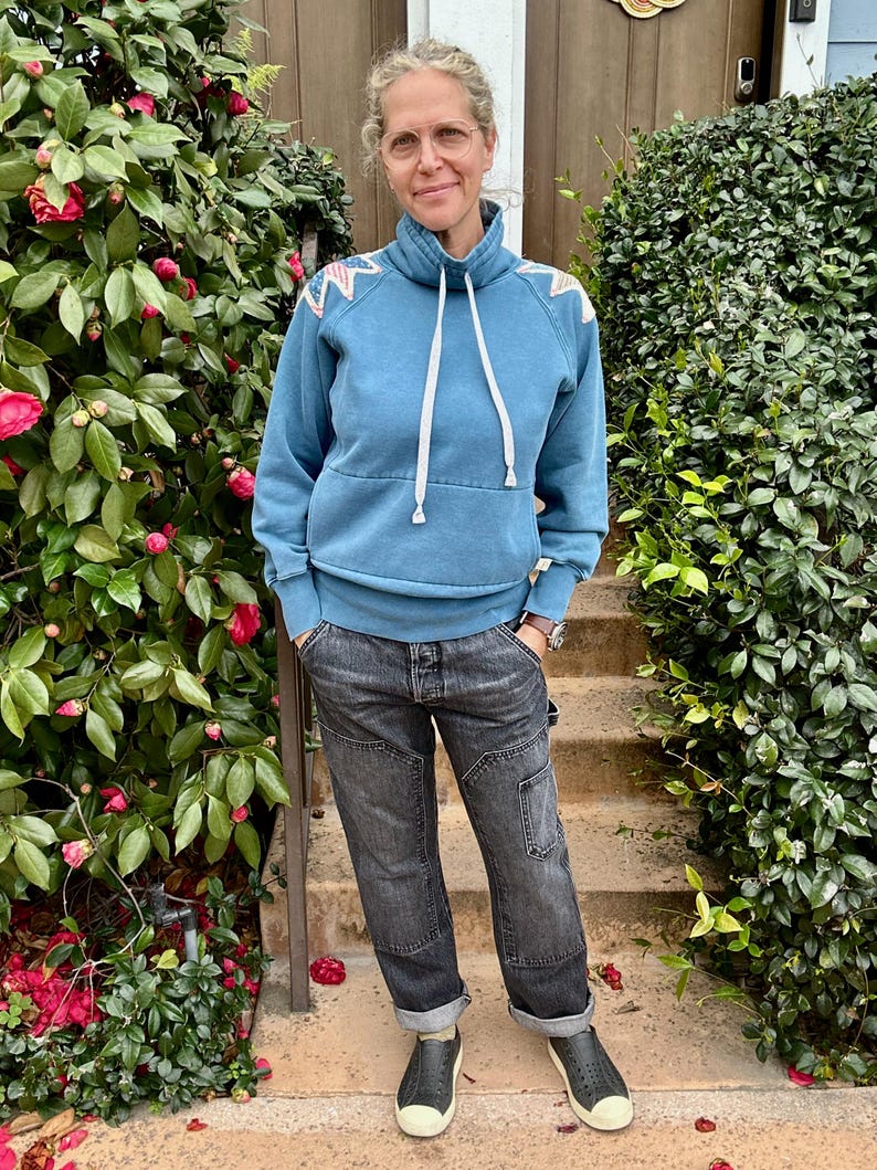 Puede incluir: Una persona con una sudadera azul con cordones blancos y cuello alto, combinada con vaqueros negros y zapatos negros. La sudadera tiene un estampado decorativo en los hombros. La persona est&aacute; de pie frente a un arbusto verde con flores rojas.