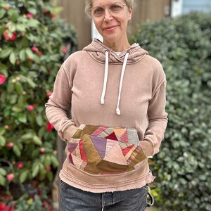 Puede incluir: Una sudadera con capucha marr&oacute;n con un bolsillo de retazos, cordones blancos y bordes desgastados. El bolsillo presenta varios patrones de tela en tonos de rojo, morado y marr&oacute;n. El modelo lleva vaqueros grises oscuros.