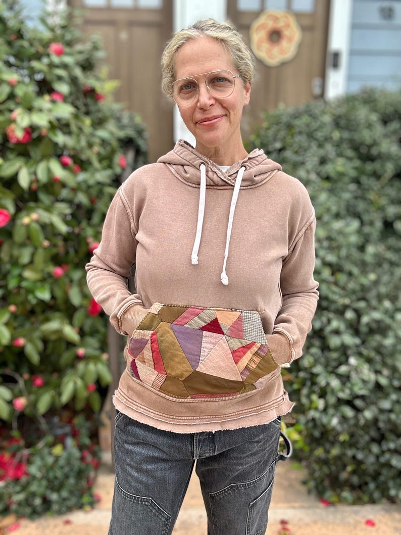 Puede incluir: Una sudadera con capucha marr&oacute;n con un bolsillo de retazos, cordones blancos y bordes desgastados. El bolsillo presenta varios patrones de tela en tonos de rojo, morado y marr&oacute;n. El modelo lleva vaqueros grises oscuros.