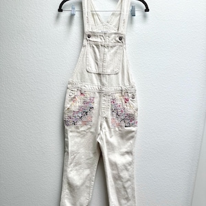 Può includere: Salopette in denim bianco sporco con una tasca sul petto e due tasche anteriori caratterizzate da un rivestimento in tessuto con motivi floreali e astratti multicolori. Il capo presenta spallacci regolabili con fibbie in metallo.