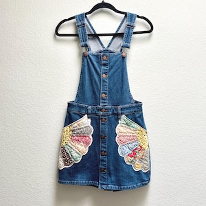 Può includere: Un vestito salopette in denim blu con chiusura frontale a bottoni e due grandi tasche a forma di ventaglio. Le tasche sono realizzate con toppe di tessuto trapuntato multicolore con vari motivi floreali e geometrici e rifinite con un bordo bianco.