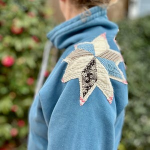 Puede incluir: Una sudadera azul claro con un gran dise&ntilde;o de estrella de retazos en el hombro. La estrella est&aacute; hecha de varias telas estampadas en blanco, negro y azul, con costuras rosas. La sudadera tiene una capucha con cord&oacute;n.