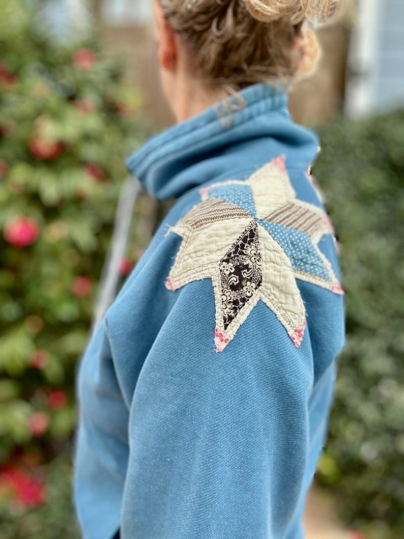 Puede incluir: Una sudadera azul claro con un gran dise&ntilde;o de estrella de retazos en el hombro. La estrella est&aacute; hecha de varias telas estampadas en blanco, negro y azul, con costuras rosas. La sudadera tiene una capucha con cord&oacute;n.