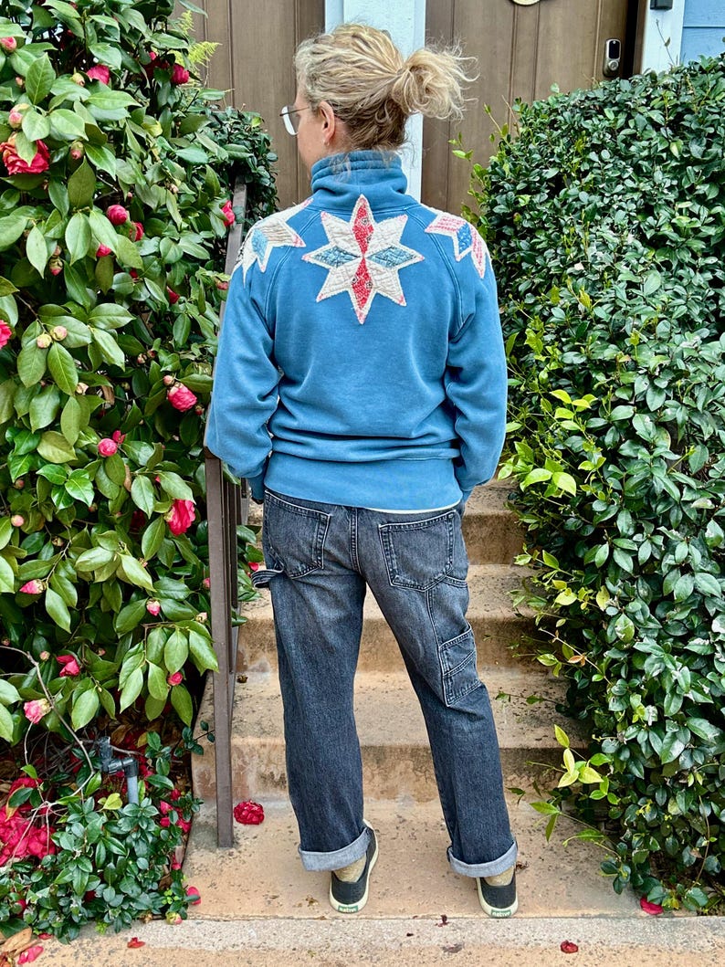 Puede incluir: Una sudadera azul con un dise&ntilde;o de estrella en la espalda, combinada con vaqueros oscuros y zapatillas negras. El dise&ntilde;o de estrella tiene detalles en blanco, rojo y azul. La persona est&aacute; de pie en escalones de hormig&oacute;n con vegetaci&oacute;n a ambos lados.