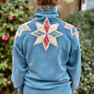 Puede incluir: Una sudadera azul con cuello alto y un parche grande en forma de estrella en la espalda. La estrella est&aacute; hecha de varias piezas de tela en rojo, blanco y azul. Parches adicionales est&aacute;n en los hombros. La sudadera es de color azul liso.
