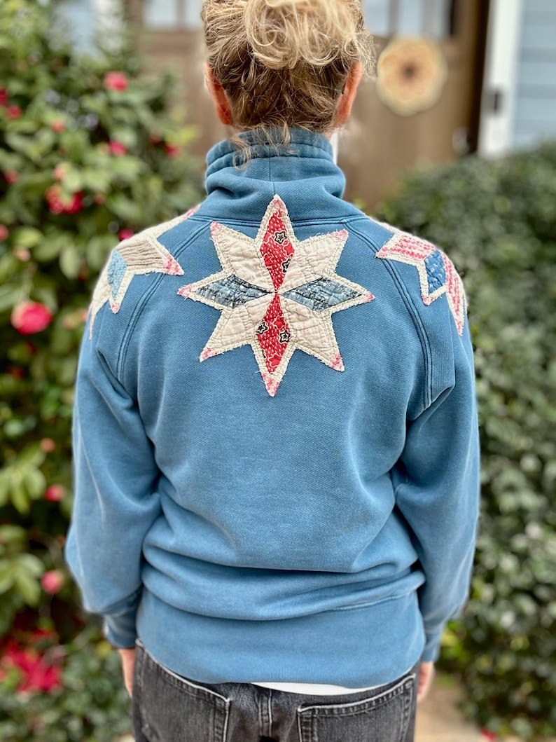 Puede incluir: Una sudadera azul con cuello alto y un parche grande en forma de estrella en la espalda. La estrella est&aacute; hecha de varias piezas de tela en rojo, blanco y azul. Parches adicionales est&aacute;n en los hombros. La sudadera es de color azul liso.