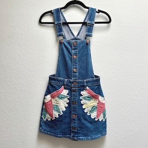 Può includere: Un vestito salopette in denim blu con chiusura frontale a bottoni e spalline regolabili. La gonna presenta due tasche anteriori decorate con toppe trapuntate a forma di ventaglio nei toni del rosa, giallo e verde acqua su fondo bianco.