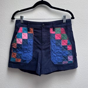 May include: Dark navy blue shorts featuring two large front pockets with a quilted patchwork design. The patchwork consists of small squares in shades of pink, blue, green, and orange, stitched together with visible thread patterns on the fabric.