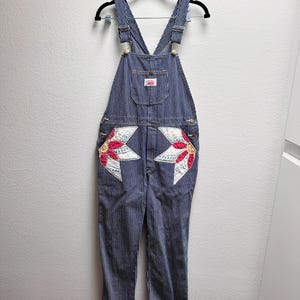 Può includere: Salopette in denim a righe hickory blu e bianche con una tasca sul petto con una piccola etichetta bianca e due grandi tasche anteriori decorate con applicazioni trapuntate a forma di stella in tessuto rosso, bianco e giallo con cuciture visibili.
