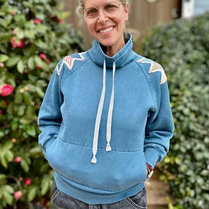 Puede incluir: Una sudadera azul con cuello alto y cordones blancos. Los hombros est&aacute;n decorados con parches en forma de estrella en rojo, blanco y azul. La persona lleva gafas y pantalones cortos vaqueros.