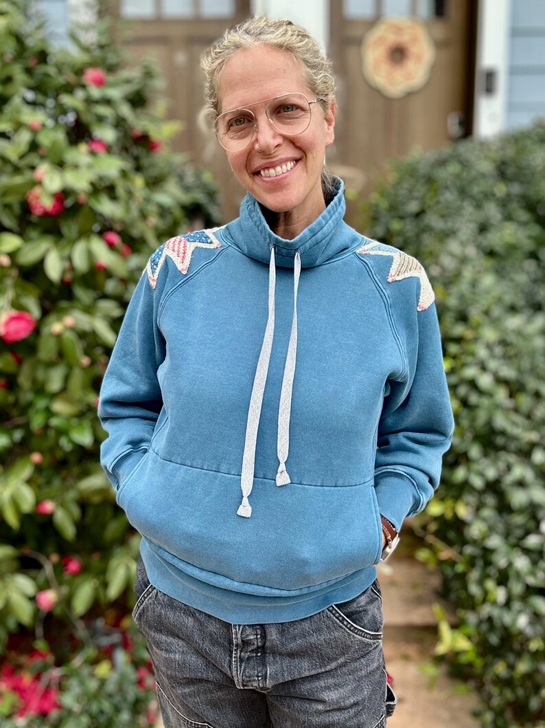 Puede incluir: Una sudadera azul con cuello alto y cordones blancos. Los hombros est&aacute;n decorados con parches en forma de estrella en rojo, blanco y azul. La persona lleva gafas y pantalones cortos vaqueros.