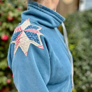 Puede incluir: Sudadera azul claro con cuello alto y parche en forma de estrella en el hombro. El parche presenta un dise&ntilde;o de patchwork con tela roja, blanca y azul, que recuerda a un edred&oacute;n.