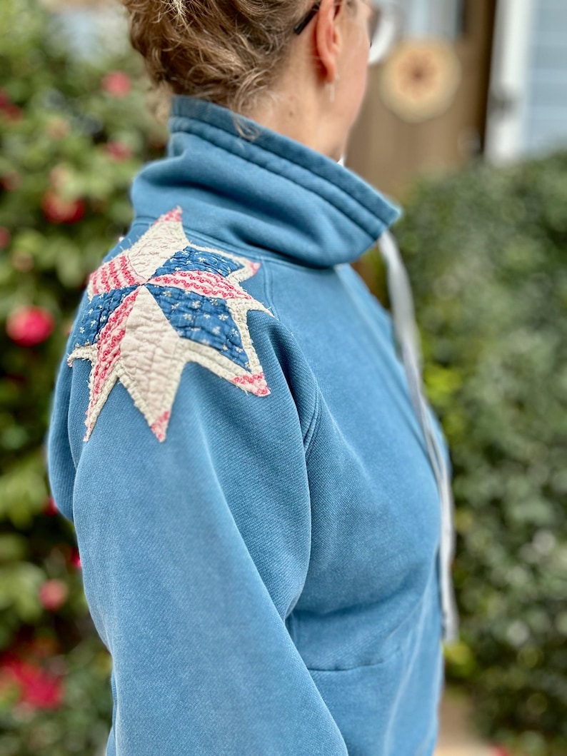 Puede incluir: Sudadera azul claro con cuello alto y parche en forma de estrella en el hombro. El parche presenta un dise&ntilde;o de patchwork con tela roja, blanca y azul, que recuerda a un edred&oacute;n.