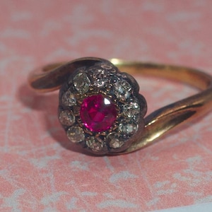 May include: A gold ring with a central, vibrant red gemstone surrounded by a halo of smaller, clear stones. The ring's band curves gracefully, creating an elegant and classic design. The ring is set on a pink surface.