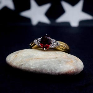 May include: A gold ring featuring a central, deep red gemstone, complemented by smaller, clear stones. The ring rests on a smooth, pale stone, set against a dark backdrop with white star shapes.