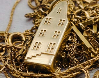 May include: A gold pendant shaped like a tall, narrow house with a gabled roof, featuring engraved rectangular windows and a small staircase detail at the base, resting on a tangled gold chain with a polished, metallic finish throughout.