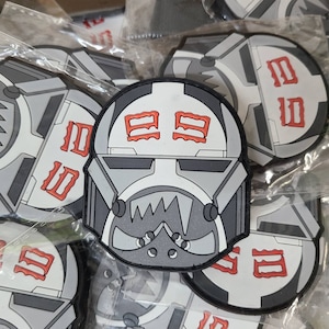 May include: A grey and white patch with a stylised helmet design and red markings. The helmet has a jagged mouth and a visor. The patch has the text "1111" on it.