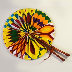 K&ouml;nnte beinhalten: Ein farbenfroher F&auml;cher mit einem gelben, orangefarbenen, roten, gr&uuml;nen, blauen und violetten Muster. Der F&auml;cher ist aus Stoff und hat einen Holzgriff.