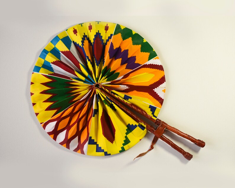 K&ouml;nnte beinhalten: Ein farbenfroher F&auml;cher mit einem gelben, orangefarbenen, roten, gr&uuml;nen, blauen und violetten Muster. Der F&auml;cher ist aus Stoff und hat einen Holzgriff.