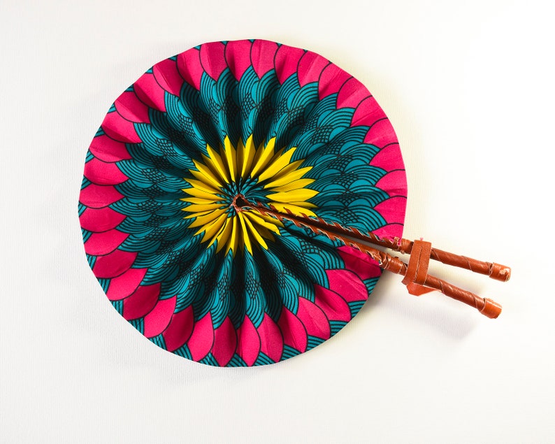 K&ouml;nnte beinhalten: Ein rosa, blauer und gelber Handf&auml;cher mit einem Holzgriff. Der F&auml;cher ist zusammengefaltet und hat eine runde Form.