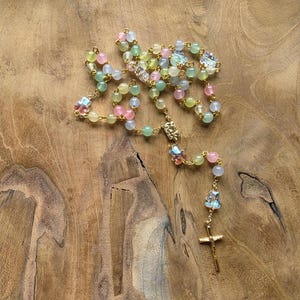 May include: A rosary featuring translucent, multi-colored beads in shades of pink, green, yellow, and blue, interspersed with small iridescent bear-shaped charms. The beads are connected by a gold-toned metal chain, ending in a gold-toned crucifix.