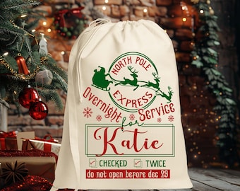 Pode incluir: Um saco de Papai Noel de lona branca com texto vermelho e verde que diz "North Pole Express Overnight Service for Katie" e "Checked Twice do not open before dec 29".