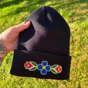 Può includere: Berretto nero con un design di ricamo floreale colorato. I fiori ricamati includono dettagli blu, rossi, gialli e verdi. Il cappello è realizzato in un morbido materiale a maglia, perfetto per il freddo.