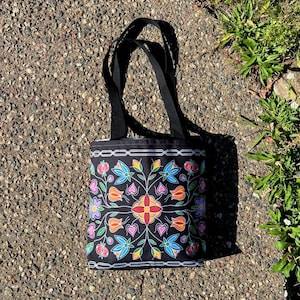 Peut inclure: Sac fourre-tout noir avec un motif floral coloré. Le motif présente des fleurs, des feuilles et des cœurs rouges, oranges, bleus, violets et jaunes. Le sac a des poignées noires et une bordure blanche. Idéal pour les courses.