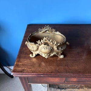 May include: An antique brass planter, featuring elaborate floral and scroll designs. The rectangular planter has a curved top and rests on four small feet. The brass has a warm, aged patina, giving it a vintage appearance. A decorative piece.